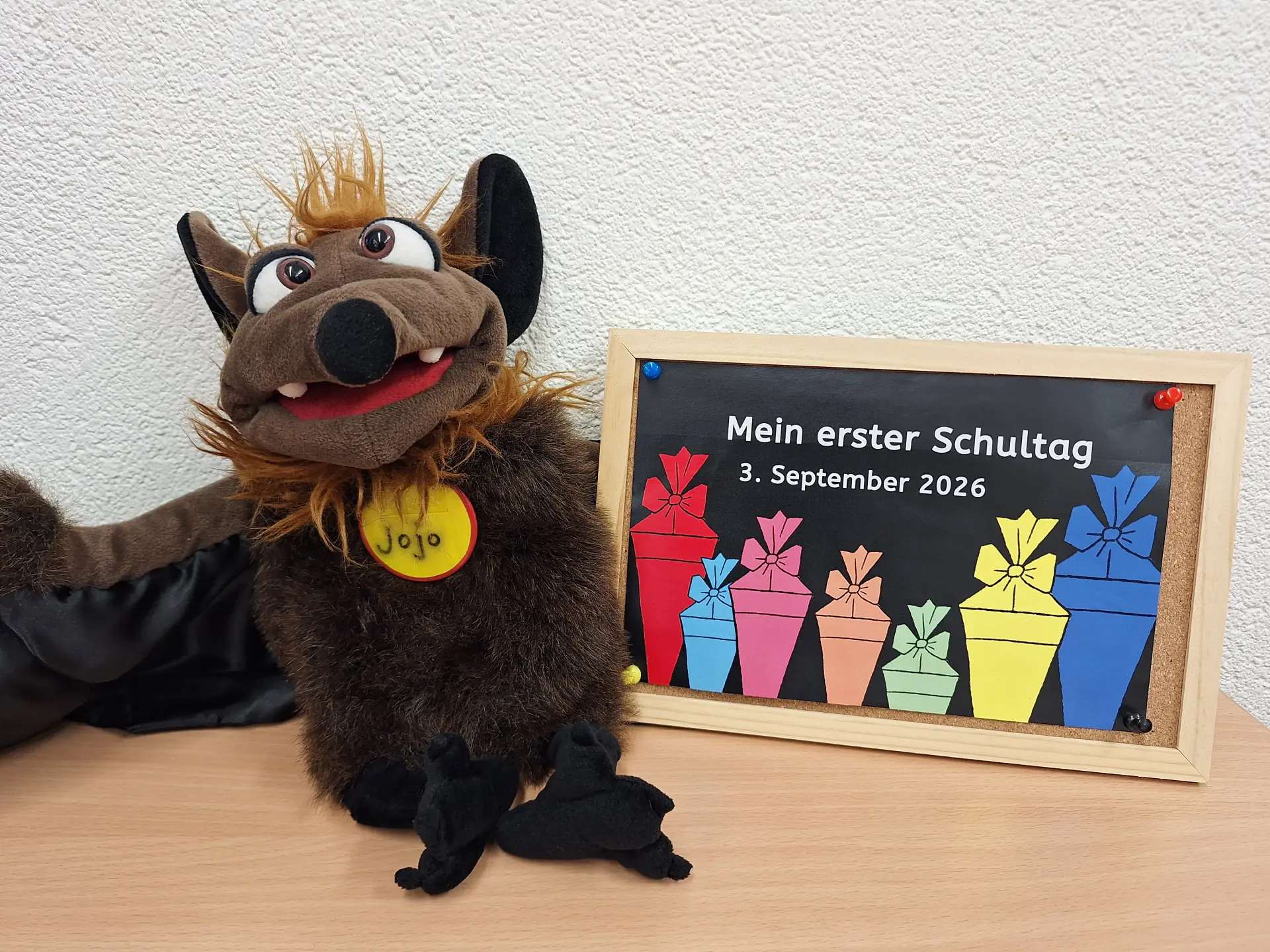 Auf dem Bild ist die Stoff-Fledermaus Jojo zu sehen, die neben einem Bild mit dem Text "Mein erster Schultag 3. September 2026" sitzt