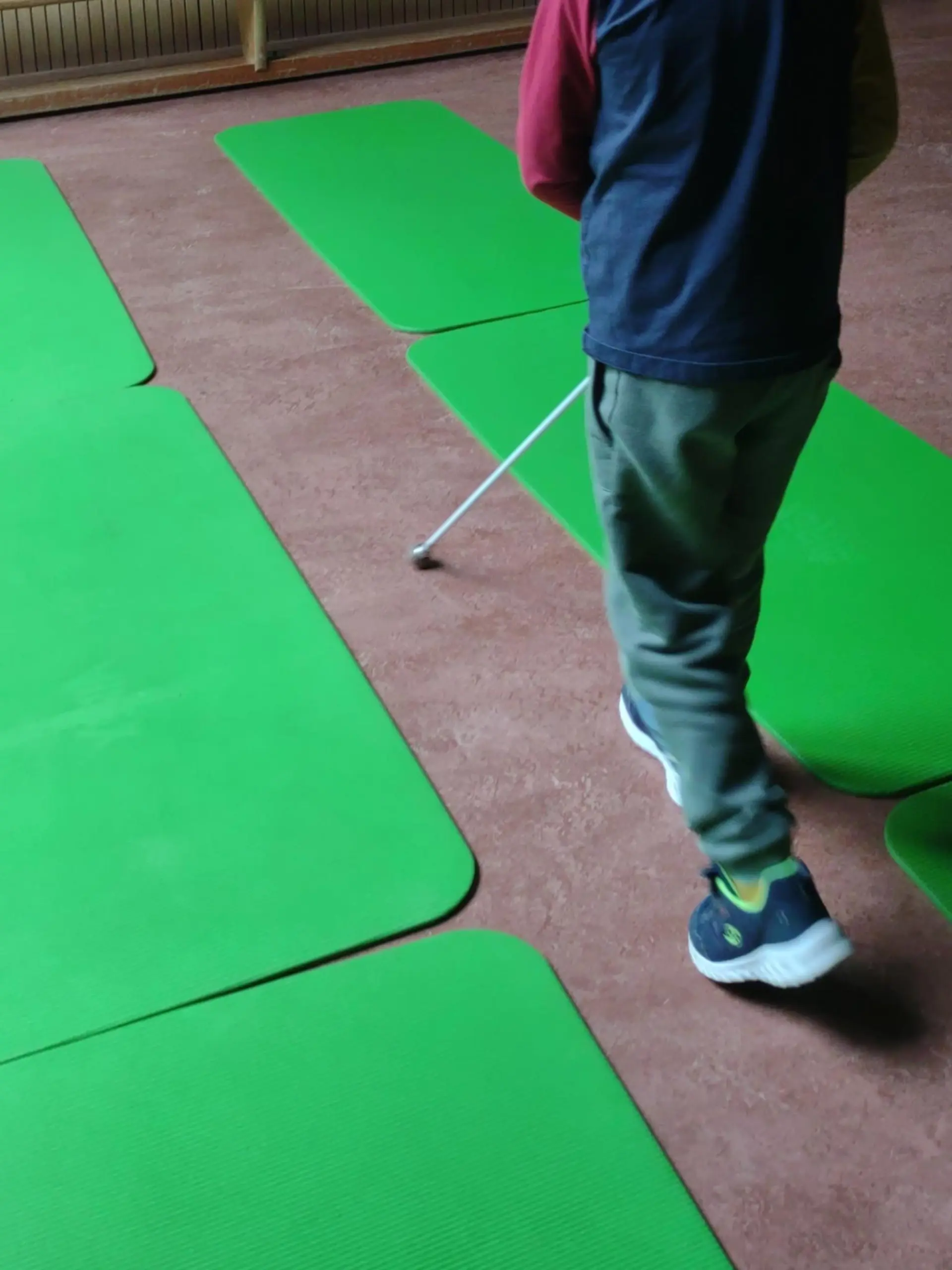 Kind ertastet mit Blindenstock Fußboden. Grüne Matten dienen als Hindernisse